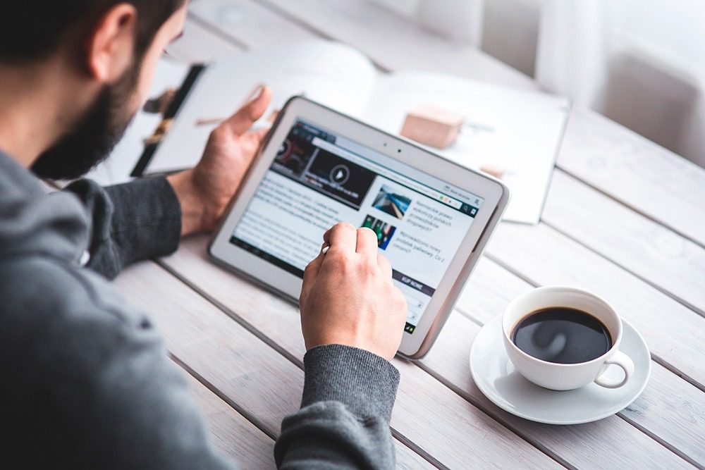 A man with a beard working on an ipad at a table with a cup of coffee to the side